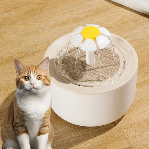 Cat sitting next to a pet water fountain with a daisy design on a wooden floor.