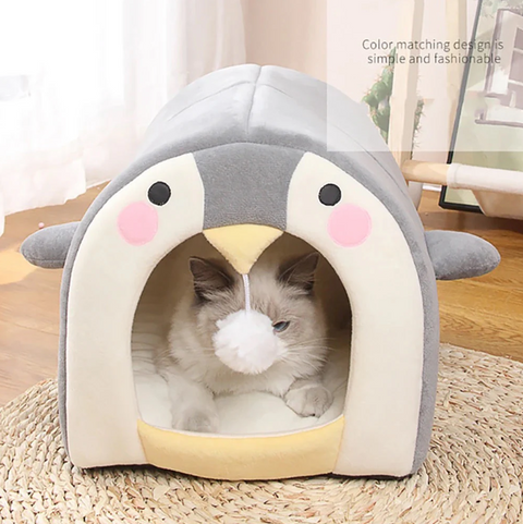 Cat lying inside a penguin-shaped pet bed on a wooden floor.